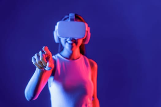 Smart female stand with surrounded by cyberpunk neon light wear VR headset connecting metaverse, futuristic cyberspace community technology. Woman using finger pointing virtual object. Hallucination.