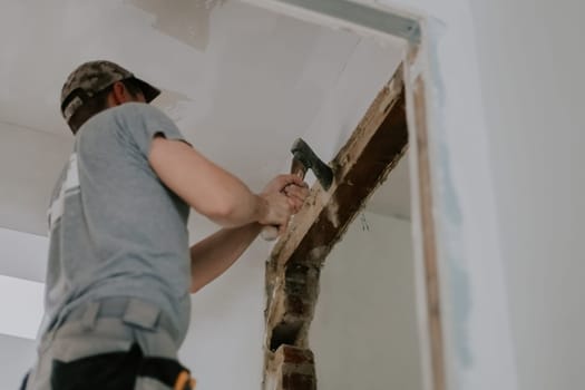 One young Caucasian man in a gray T-shirt and cap clears old putty from the top of a doorway with an ax while standing on a stepladder, close-up view from below with selective focus. Construction work concept.