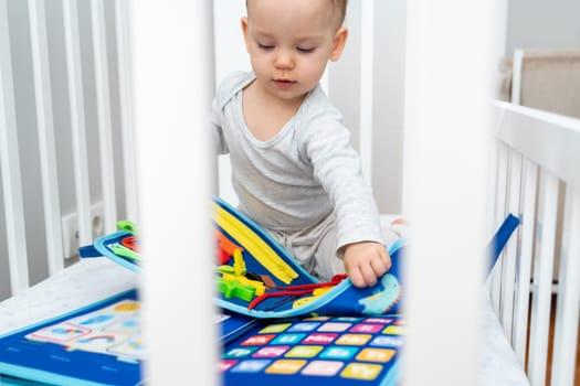 Little baby girl playing with busy book while sitting in crib. Concept of smart quiet books and modern educational toys