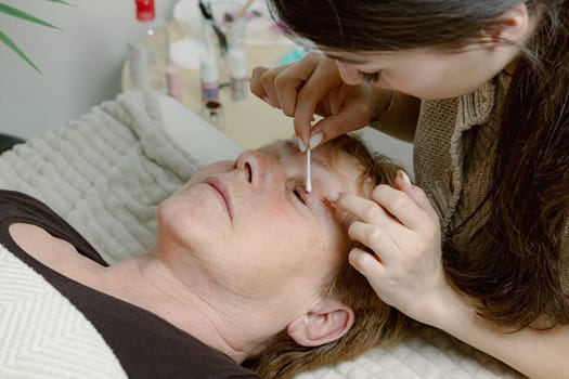 One Caucasian young beautiful brunette cosmetologist degreases the upper eyelid of the eye with a cotton swab to an elderly female client preparing them for the lamination procedure, which is lying on the cosmetology table in a home beauty salon, side view close-up.