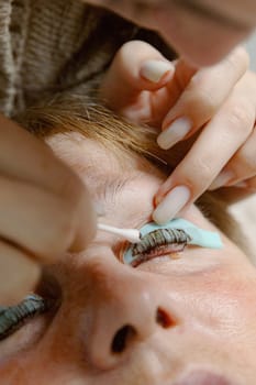 One Caucasian young beautiful brunette cosmetologist wipes excess brown paint with a cotton stick from the eyelash of the left eye with a sponge stick to an elderly female client who is lying on the cosmetology table in a home beauty salon, side view very close-up.