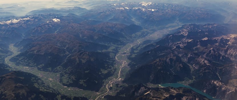 Beautiful panoramic view of the rocky dark tops of the Alpine mountains in places with snow, settlements and country roads from the window of a flying airplane over Switzerland, close-up side view.