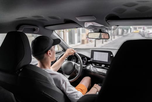 A beautiful view of one young Caucasian guy in a cap sitting half sideways behind the wheel of a car driving along the road on a summer day, reflected in the rearview mirror, close-up side view from above.