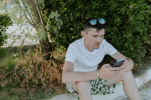 A portrait of one young Caucasian guy with sunglasses on his head sits on the curb near the bushes in a city park, enthusiastically communicates on a smartphone on a sunny summer day, close-up side view.