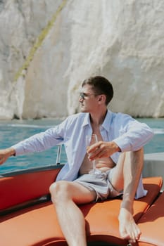 Portrait of one young Caucasian handsome guy in sunglasses sits at the stern of a boat, holds a glass of champagne in his hand, looks away and enjoys sailing on the sea on a private boat on a sunny summer day, close-up side view with selective focus.