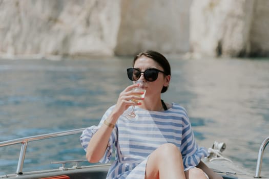 Portrait of one young Caucasian beautiful girl in sunglasses sitting in a boat and happily drinking from glasses of champagne against the background of blurred rocks while sailing on the sea on a sunny summer day, close-up side view.