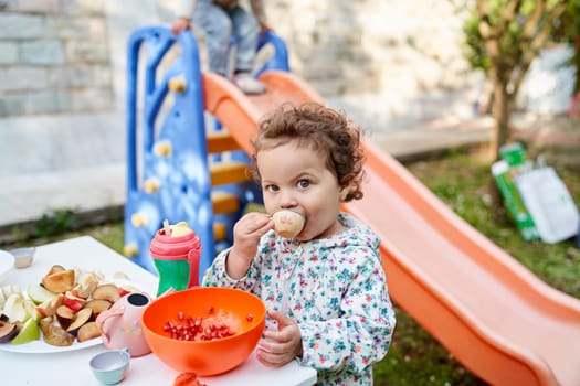 Little girl eats pomegranate seeds from a small cup while standing at a festive table in the garden. High quality photo