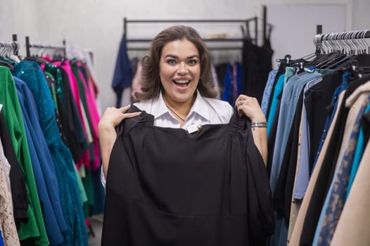 A fat woman in a plus size store tries on a black evening dress. Vertical photo