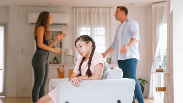 Annoyed and unhappy young girl sitting on sofa trapped in middle of tension by her parent argument in living room. Unhealthy domestic lifestyle and traumatic childhood develop to depression Synchronos