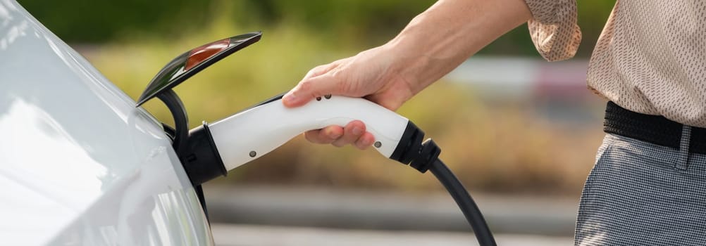 Young man recharge electric car's battery from charging station in city commercial parking lot. Rechargeable EV car for sustainable environmental friendly urban travel. Panorama Expedient