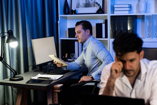 Colleagues concentrating on their job task at night home office behind desk while another man with thoughtful face solving to postpone the deadline of project at the blurred front side. Sellable.