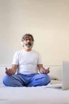 Vertical portrait of mature adult man sitting on bed at home following online meditation classes using laptop and headphones. Male meditating in the morning. Mental health and technology concept.