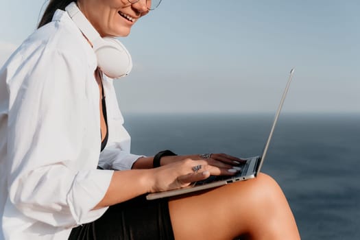 Digital nomad, woman in the hat, a business woman with a laptop sits on the rocks by the sea during sunset, makes a business transaction online from a distance. Freelance, remote work on vacation.