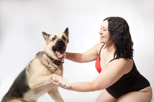 Portrait of attractive thick woman in red black swimsuit with big dog shepherd on white background. Body positive, selfie of Funny plus size model