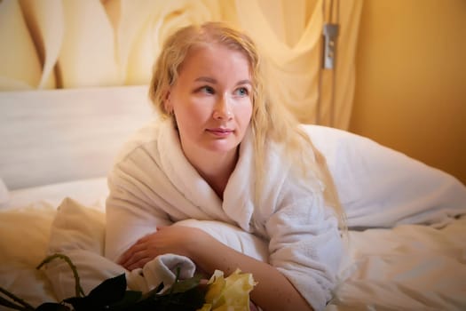 A blonde girl in a white terry bathrobe in hotel room with flowers. Wedding travel, Weekend and vacation, body care