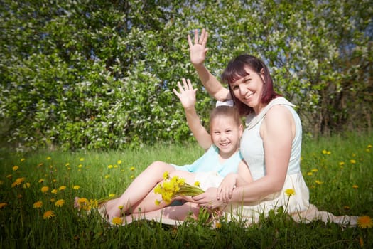 Happy mother and daughter enjoying rest, playing and fun on nature on a green lawn with dandelions and blooming apple tree on background. Woman and girl resting outdoors in summer and spring day