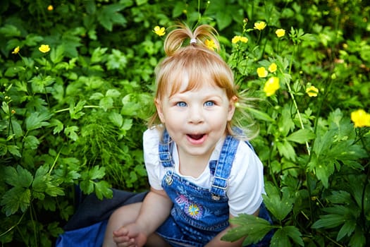 Joyful Toddler Among Spring Flowers. Toddler with bright blue eyes sits among blooming yellow flowers, smiling. A healthy child in the fresh air. Good ecology