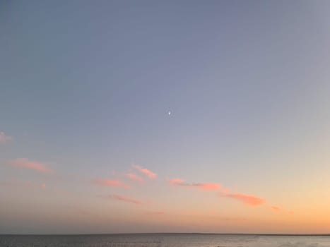 Little moon hangs in the sky over the ocean during dusk, accompanied by a vibrant afterglow. A beautiful natural landscape at sunset.