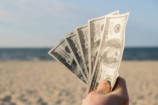 Unrecognizable woman Displaying Spread of Cash dollars bills on sandy beach coastline. Concept finance saving money for holiday vacation. Costs in travel holidays. Extra money, passive income