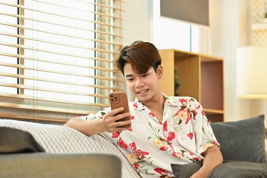 Smiling man sitting on floor in living room and using mobile phone.