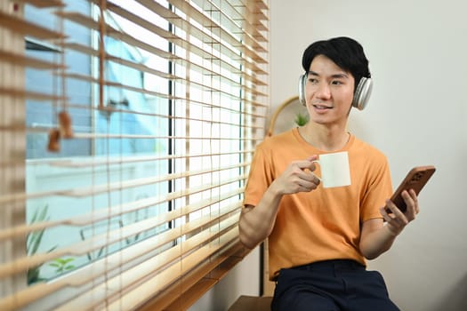 Carefree young asian man sitting by the window with a cup of coffee and looking away.