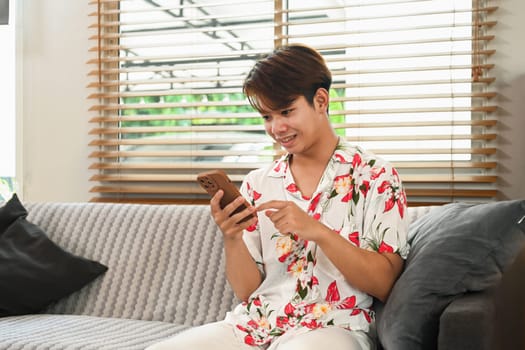 Pleased young Asian man typing messages on mobile phone sitting on sofa in living room.