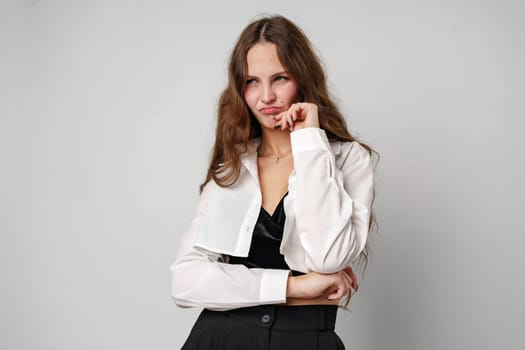 A young woman stands against a neutral gray backdrop, wearing a white jacket and black attire. She is gazing to the side with a thoughtful, pensive expression, her hand resting thoughtfully on her chin, as if deep in contemplation or making a decision.