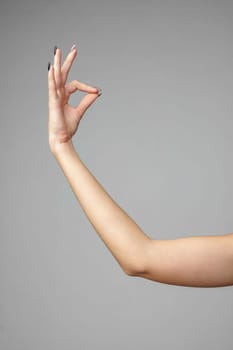 A close-up view captures a persons arm extended from the left with their fingers forming the okay sign, consisting of a circle made by the thumb touching the index finger, with the background in a neutral gray tone.