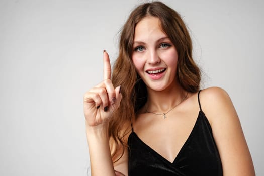 A smiling young woman with long wavy hair raises her index finger as if shes about to express an idea. She wears a black velvet dress and has a playful look on her face. The light grey background provides a neutral backdrop that highlights her figure.