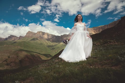 a bride in a white wedding dress walks in the green mountains on the grass, in the background there are mountains and a lake.