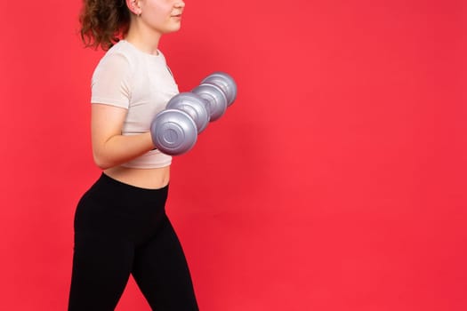 Teenage sportive girl exercises with dumbbells to develop muscles isolated on a red background.