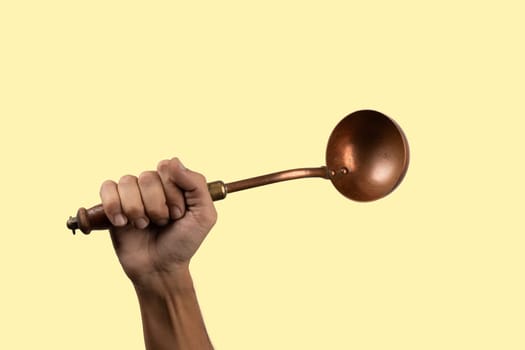 Black male hand holding a brass vintage kitchen ladle isolated on yellow background. High quality photo