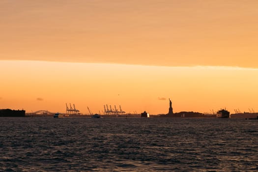 Scenic image capturing the Statue of Liberty with a beautiful sunset backdrop over the New York City harbor. The sky is lit with warm hues, and boats are visible on the water.