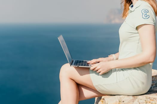 Digital nomad, woman in the hat, a business woman with a laptop sits on the rocks by the sea during sunset, makes a business transaction online from a distance. Freelance, remote work on vacation.