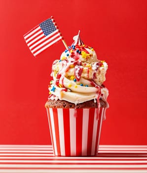 Cupcake with ice cream topped with American flag, colorful sprinkles, cream swirls, on red and white striped background.