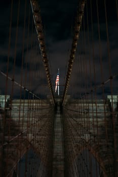 A captivating view of the Brooklyn Bridge at night featuring an American flag illuminated at the center. The iconic structure is showcased against a dark sky, highlighting its architectural beauty.