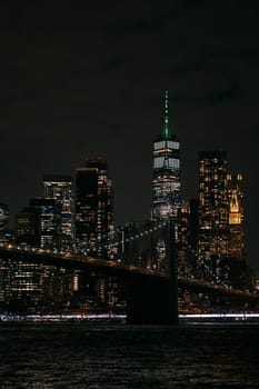 A stunning night view of the New York City skyline featuring the iconic Brooklyn Bridge and brightly lit skyscrapers. The image captures the vibrant, bustling atmosphere of the city after dark.
