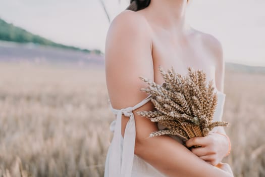 A woman is holding a bunch of wheat in her arms. The wheat is dry and brown, and the woman is wearing a white dress. The scene is set in a field, and the woman is posing for a photo