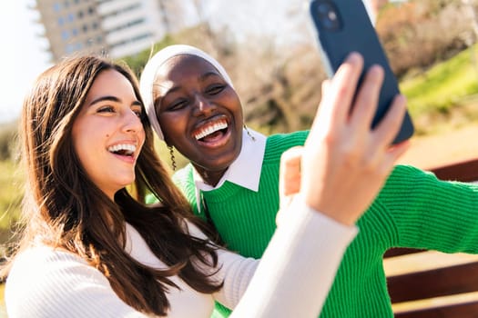 two young female friends laughing happy and having fun taking selfie photo with mobile phone in a city park, concept of diversity and modern lifestyle