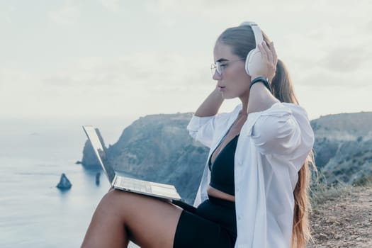 Digital nomad, woman in the hat, a business woman with a laptop sits on the rocks by the sea during sunset, makes a business transaction online from a distance. Freelance, remote work on vacation.