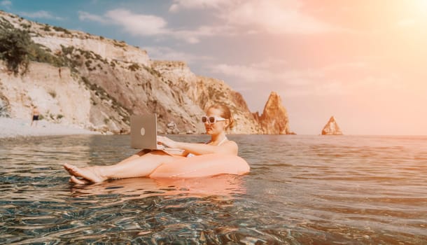 Woman freelancer works on laptop swimming in sea on pink inflatable ring. Pretty lady typing on computer while floating in the sea on inflatable donut at sunset. Freelance, remote work on vacation