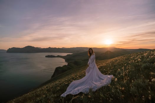 A woman stands on a hill overlooking a body of water. The sky is a mix of blue and orange, and the sun is setting. The woman is wearing a long white dress, and she is enjoying the view