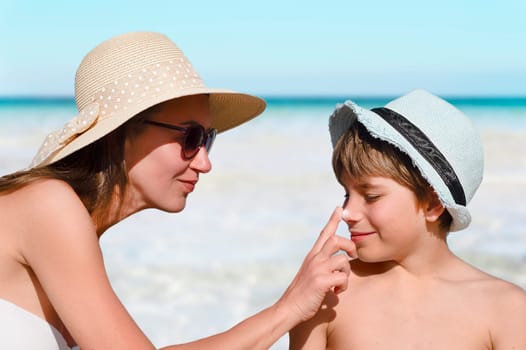 Woman carefully smears her child's face with protective cream on the beach against background of a clear sea. Skin care. Protection from the sun. Sunscreen for children.