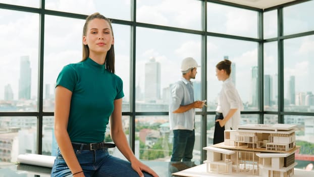 Young beautiful interior designer looking at camera while engineer team talking about house design. Skilled businesswoman smiling at camera with confident while sitting near house model. Tracery