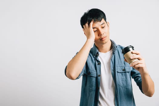 A tired and sad Asian man, eyes closed, holds a coffee cup, battling exhaustion. Studio shot isolated on white, conveying the weight of overwork. Copyspace included.