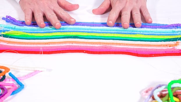 Hands touch a neatly organized rainbow of colorful, coiled clay bead strands against a contrasting white surface.