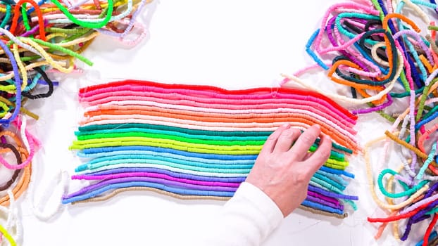 Flat lay. Hands touch a neatly organized rainbow of colorful, coiled clay bead strands against a contrasting white surface.