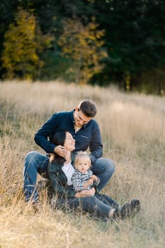 Mom with a little girl on her knees sits on the lawn leaning against dad sitting behind her. High quality photo