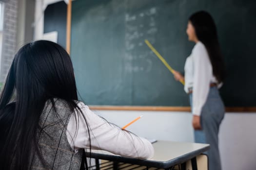 A woman is teaching a child in a classroom. The child is sitting at a desk with a pencil and a piece of paper. The woman is standing in front of a blackboard with a chalk