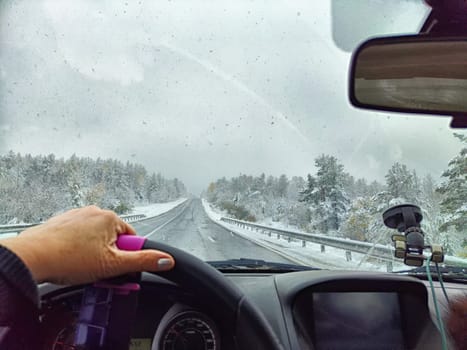 Car salon, windshield, hand on steering wheel and landscape with snow in cloudy weather. Beginning of winter, slippery road. Partial focus. Blurred